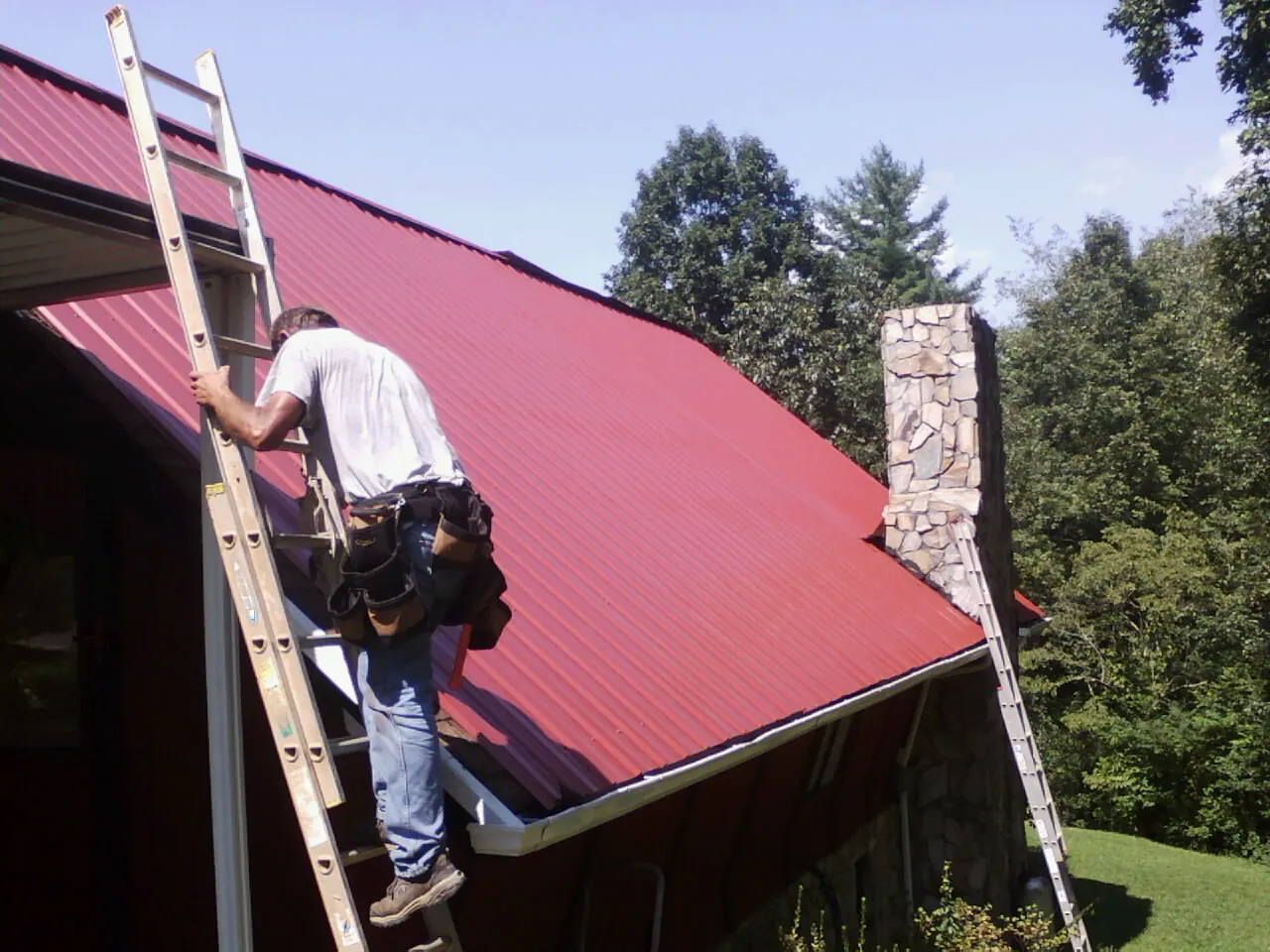 Roofer on a ladder beside a red metal roof and stone chimney — Soffit & Fascia Repair by Roofing Brothers in North Woodstock